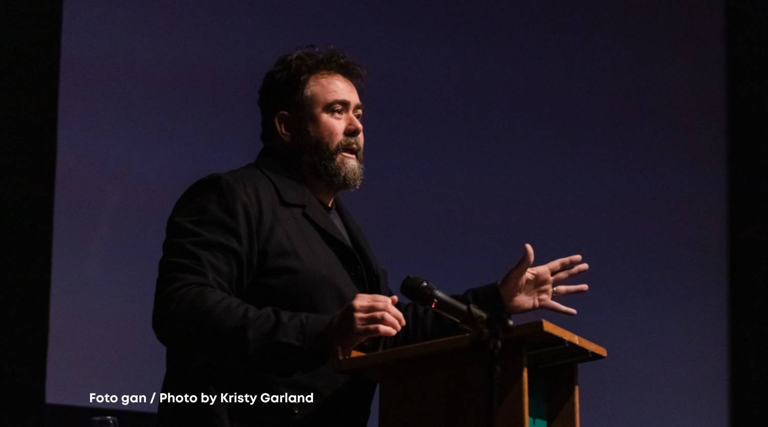 Celyn Jones stands against a dark blue background, at a lectern. He's white, with dark curly hair and wearing a dark suit